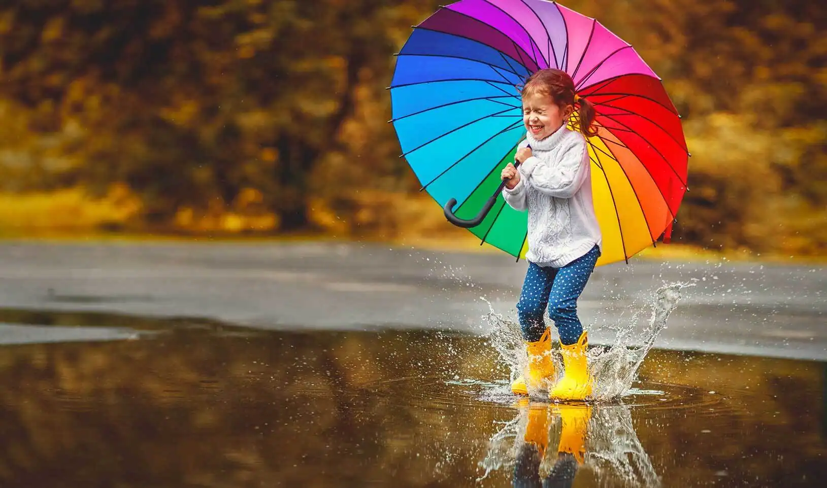 A small girl happily stomping on a puddle while holding a rainbow umbrella