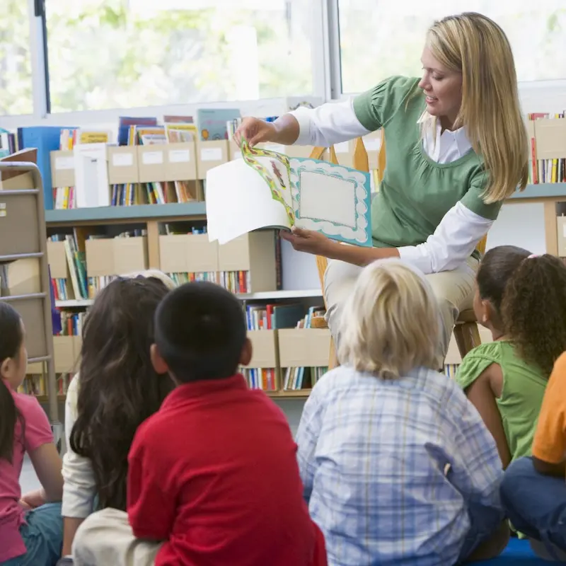 A teacher reading through a book in front of a row of children sat on the floor